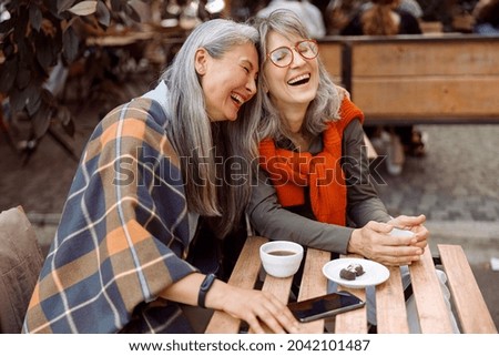 Pretty senior Asian woman puts head on laughing friend shoulder resting together at table in street cafe on nice autumn day Royalty-Free Stock Photo #2042101487