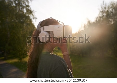 The girl holds virtual reality glasses on her head with her hands, stands against the background of green trees. The concept of the future. High quality photo