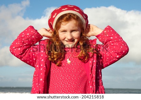 portrait of a little red headed girl in her red winter hat at a beach 