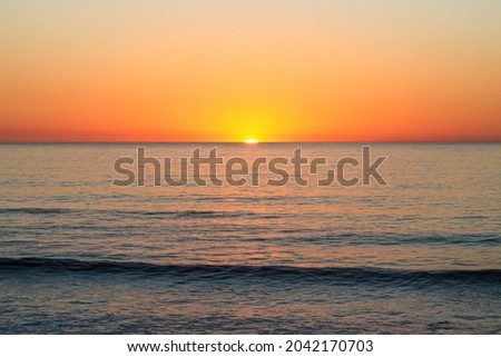 The sun nearly vanished at the horizon over a nearly calm ocean with some smaller waves visible in the foreground