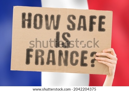 The question " How safe is France? " on a banner in men's hand with blurred French flag on the background. Safety. Street. Outdoor. Dangerous. Security. Attack. Criminal. Criminality