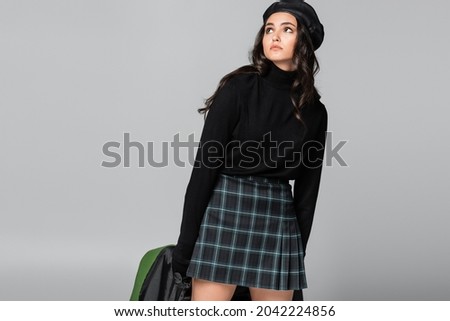 young trendy woman in beret and plaid skirt holding green leather jacket isolated on grey