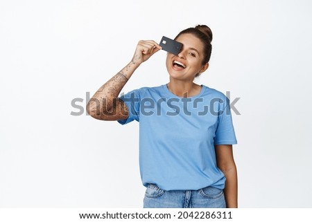 Portrait of beautiful happy woman with tattoos, showing credit card against eye, laughing and smiling, satisfied bank client, standing against white background