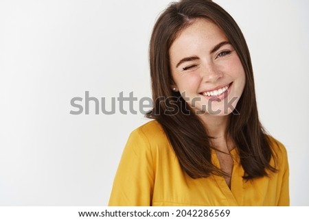 Close-up of cute and dreamy young woman smiling, winking at camera to encourage client, standing over white background Royalty-Free Stock Photo #2042286569