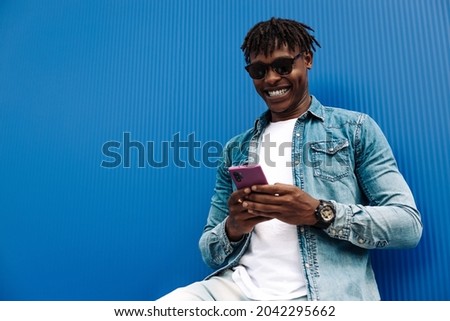 african man typing sms on blue background, 5g internet concept, high speed internet on phone, black man in blue sacker, and jeans