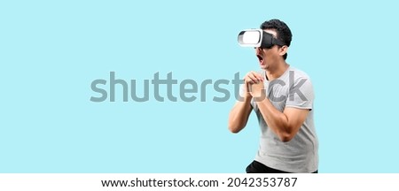 Asian man playing VR video game with virtual reality goggles and Shock and surprise face ,trying to touch something on light blue background in studio With copy space.
