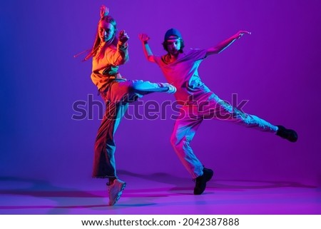 Street style. Stylish man and woman dancing hip-hop in casual youth clothes on gradient purple pink background at dance hall in neon light. Youth culture, hip-hop, fashion, breakdance, action and ad