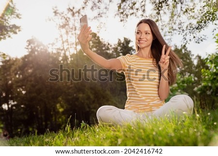 Portrait of attractive cheerful girl sitting on grass taking selfie showing v-sign good mood rest fresh air outdoors