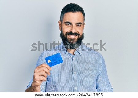 Young hispanic man holding credit card looking positive and happy standing and smiling with a confident smile showing teeth 