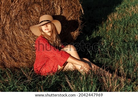 Stylish girl resting near a bale of hay in a summer field at sunset.Young blonde woman in a straw hat sits by a haystack, an atmospheric calm moment. Rural slow life