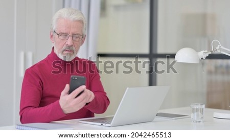 Old Man with Laptop Browsing Internet on Smartphone at Work