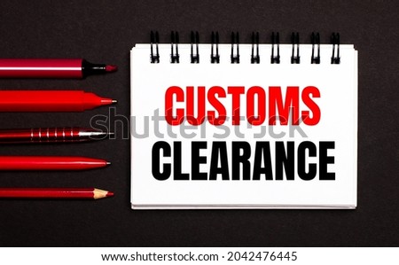 A white notebook with the text CUSTOMS CLEARANCE next to red pens, pencils and markers on a black background.