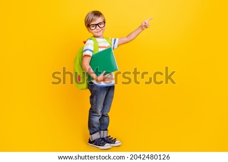 Full body photo of little clever blond boy point empty space wear white t-shirt bag jeans sneakers eyewear isolated on yellow background