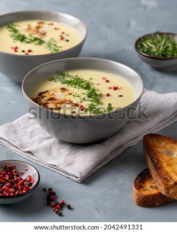 Cauliflower soup on gray tabletop, creamy flower soup with toasted bread, garden cress, red and black peppercorns.  Royalty-Free Stock Photo #2042491331