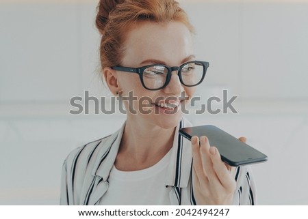 Portrait of happy cheerful red-haired woman in eyewear holding mobile phone, recording audio message and smiling while spending time at home, dressed casually, positive female talking on speakerphone