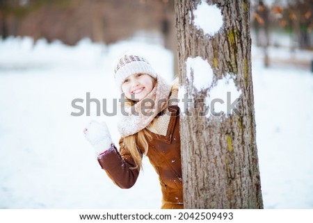 smiling stylish girl with mittens in a knitted hat and sheepskin coat playing snowball outdoors in the city park in winter.