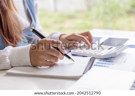 Female employee is taking notes and pressing a calculator, she is summarizing the financial documents for a meeting with the head of department, he works in the finance department. Financial concepts. Royalty-Free Stock Photo #2042512790