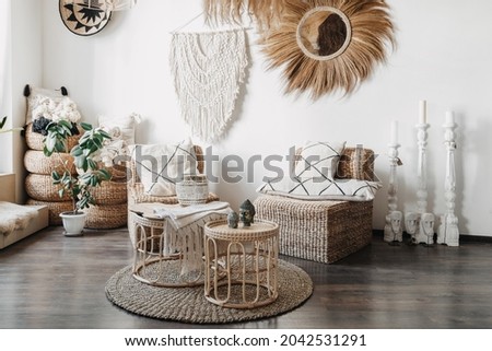 Home decor concept. Comfortable wicker furniture, rattan armchair with cushions, bamboo coffee table and macrame on white wall in cozy living room with ethnic interior design Royalty-Free Stock Photo #2042531291