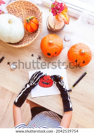 A child in black gloves with bones draws a laughing pumpkin while sitting by the window, surrounded by pumpkins and spiders, top view. Preparing for the holiday, decorating the house for Halloween.