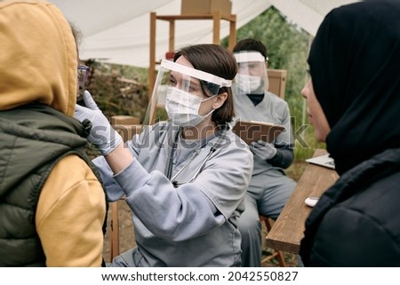 Young female medical worker in protective face shield checking throat of refugee child outdoors Royalty-Free Stock Photo #2042550827