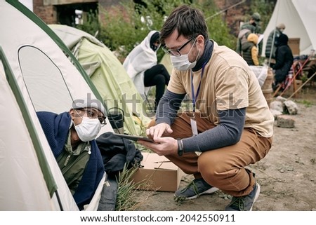 Young social worker with badge crouching at tent and making notes in tablet while talking to refugee Royalty-Free Stock Photo #2042550911