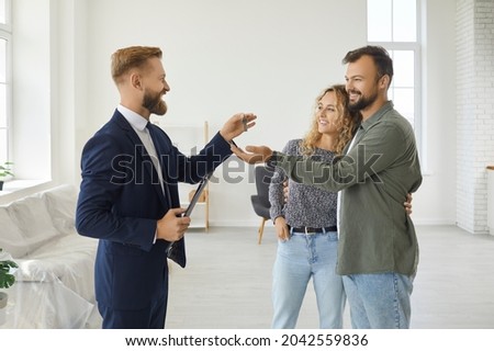 Professional realtor standing in empty living room of new apartment gives house keys to happy married couple. Young boyfriend and girlfriend or husband and wife take keys that real estate agent gives Royalty-Free Stock Photo #2042559836