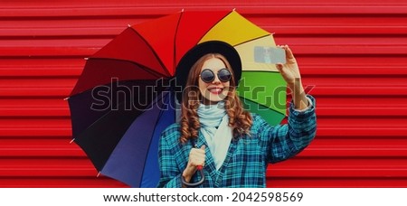 Portrait of happy smiling young woman taking a selfie picture by phone holding colorful umbrella on red background