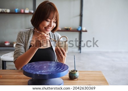 Beautiful smiling young woman attaching handle to clay mug she created and using brush and little water to smooth connections