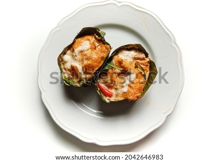 Close-up top view photo of steamed fish fillet with curry paste, coconut milk and basil in banana leaf krathong on a white plate, shot with light and shadow for decorative design and advertising.