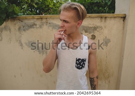 Blonde tattooed young man, wearing white t-shirt, smoking on a old wall background. City youth lifestyle, habits