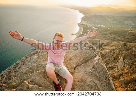 A happy man is sitting with his hands open on the edge of a cliff above the sea with a beautiful view of the mountains and the village in the Crimea. The conquest of heights and the concept of freedom