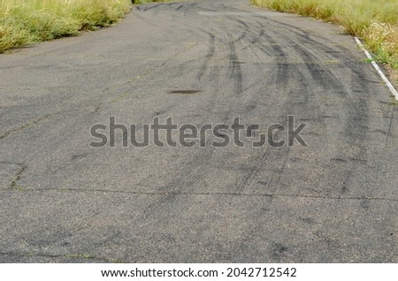 Gray asphalt with black tire tracks. An old country road with cracks. Green grass on the side of the highway. Curved wheel tracks in a bend in the driveway.