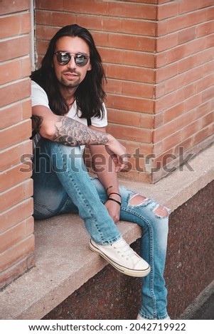 Attractive young man wearing sunglasses and long hair relaxing in urban background. Sunglasses male model with long hair and tattoos on hand.