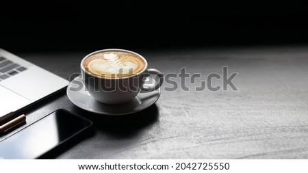 Focus on white coffe cup with latte art coffee and laptop, mobile phone on desk, Dark tone