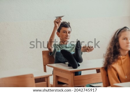 A naughty boy with his feet on a desk sits in the classroom and throws a plane. Selective focus . High quality photo Royalty-Free Stock Photo #2042745917