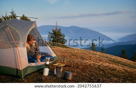 Woman traveler sitting in camp tent and taking photo of tourist gas burner and kettle. Female hiker using mobile phone while resting in tourist tent on grassy hill with mountain and sky on background.