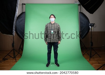 Front view of young Asian man in a gray plaid shirt and navy pants standing on a green screen background with LED softbox lights on tripods illuminate to him, get ready for a shoot in the studio.