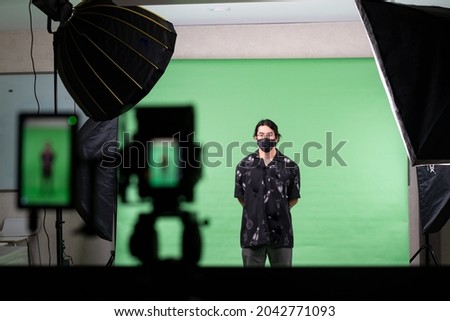 Selective focus of young Asian man wearing glasses and a black face mask, standing on a green screen background with the back of softbox light, reflector, and a blurred of camera in the foreground.