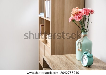 Vase with beautiful carnations and alarm clock on table in room