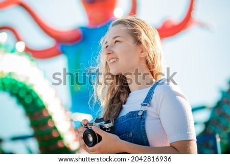A young woman tourist in a denim sundress stands with cameras in an amusement park. Teen girl in modern clothes takes pictures on the background of the carousel