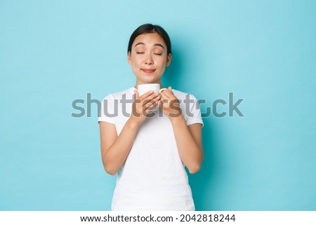 Portrait of delighted dreamy, pretty asian girl in white t-shirt, close eyes and smelling coffee, enjoying morning routine, sipping cappuccino from mug, standing light blue background Royalty-Free Stock Photo #2042818244
