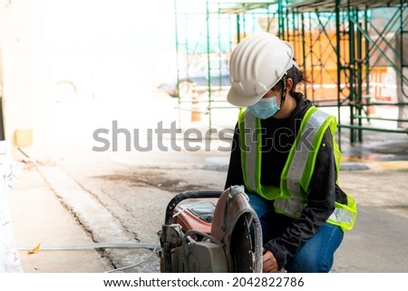 Young Asian female engineer wearing a medical mask to cover her mouth and wearing a white safety helmet A small concrete slab cutting machine is being prepared in the construction Zone.