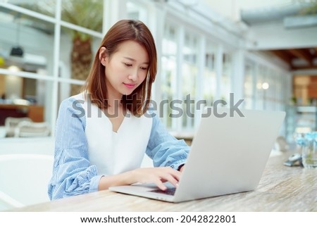 A beautiful woman wearing an Asian white shirt is sitting in front of a laptop computer shopping online with a happy smile in a bakery shop.