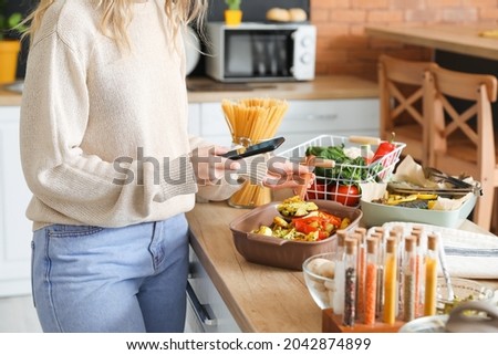Young woman taking photo of baking dish with vegetables in kitchen