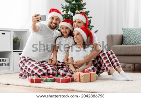 family, winter holidays and people concept - happy mother, father and two daughters in santa hats sitting under christmas tree at home and taking selfie with smartphone