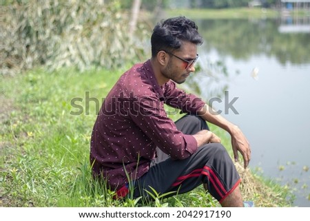 A young boy is sitting on the ground by the river wearing a brown shirt and black sunglasses with styling and his behind the background blur.