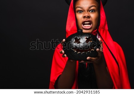 spooky african american boy in devil costume holding dark carved pumpkin isolated on black
