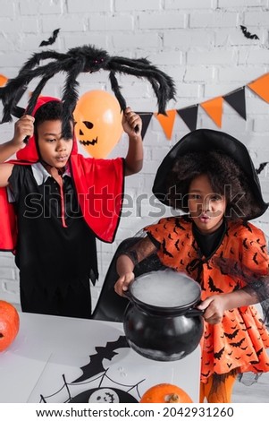 african american kid in halloween costume holding witch cauldron with potion near brother with toy spider
