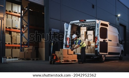 Outside of Logistics Distributions Warehouse Delivery Van: Worker Unloading Cardboard Boxes on Hand Truck, Online Orders, Purchases, E-Commerce Goods, Food, Medical Supply. Evening Shot Royalty-Free Stock Photo #2042952362