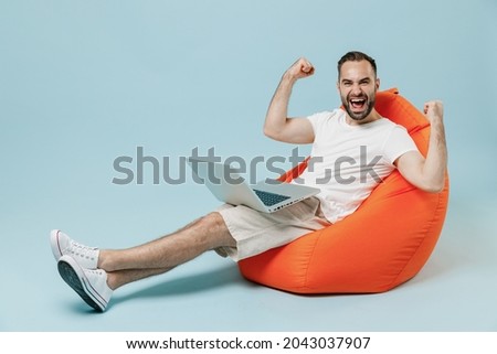 Full length young happy overjoyed man wear white t-shirt sit in bag chair hold use work on laptop pc computer rest relax do winner gesture clench fist isolated on plain pastel light blue background.